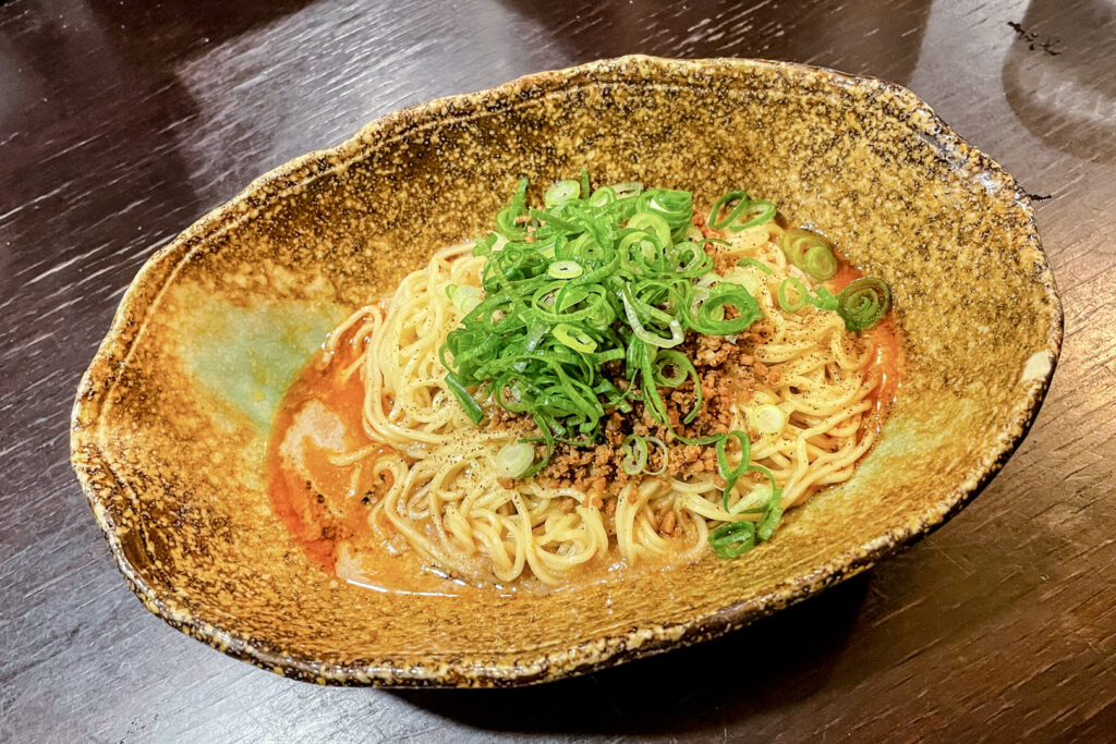 Hiroshima food: Shiru Nashi Tantanmen served in a brown-earthy toned oval ceramic bowl. The noodles are resting on a small amount of spicy broth/sauce and are topped with ground meat and lots of scallions (spring onions).