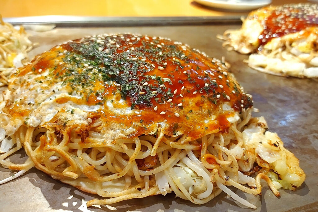 A side-on view of a Hiroshima style okonomiyaki on a hotplate. A generous filling of noodles, cabbage, meat and bean shoots can be seen between the egg and pancake, and it's all been topped with okonomiyaki sauce, aonori seaweed and some sesame seeds.
