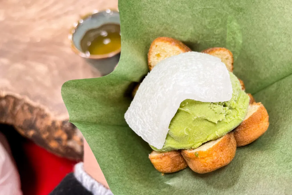 A 'momiji manju' cake cut in half but not quite all the way through, and filled with a scoop of matcha ice cream and a thin mochi rice cake on top. It has been served in a napkin (green in color) so it can be more easily held and eaten.
