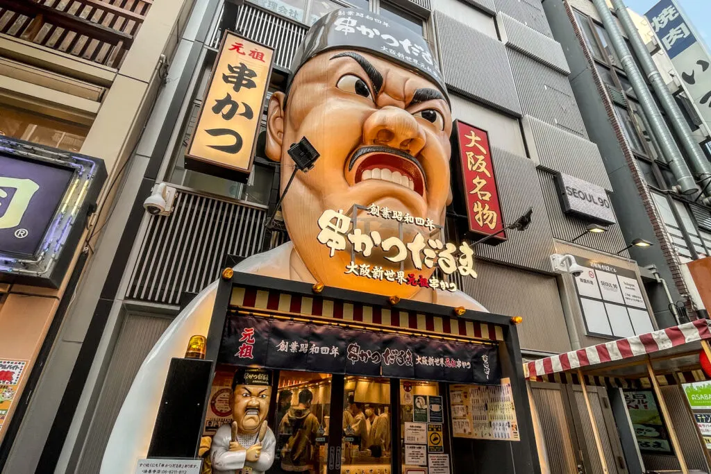 The massive face above the entrance to the Kushikatsu Daruma restaurant on Dotonbori, Osaka. The face is of a male chef with a black hat and white attire. His expression is cartoonish and over-the-top, almost a little angry-looking with furrowed brows, big flared nostrils and bared teeth. 