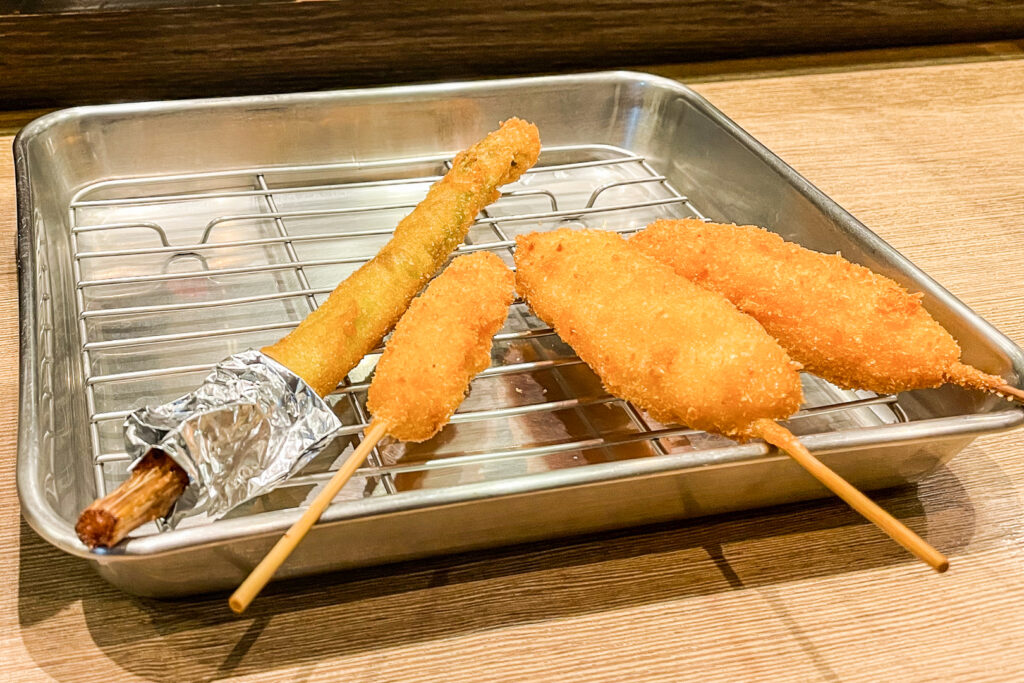 A silver rectangular metal tray with a cooling rack nested inside. Four pieces of kushikatsu are resting on top. From right to left: Pork, chicken tender, mozzarella and asparagus. 