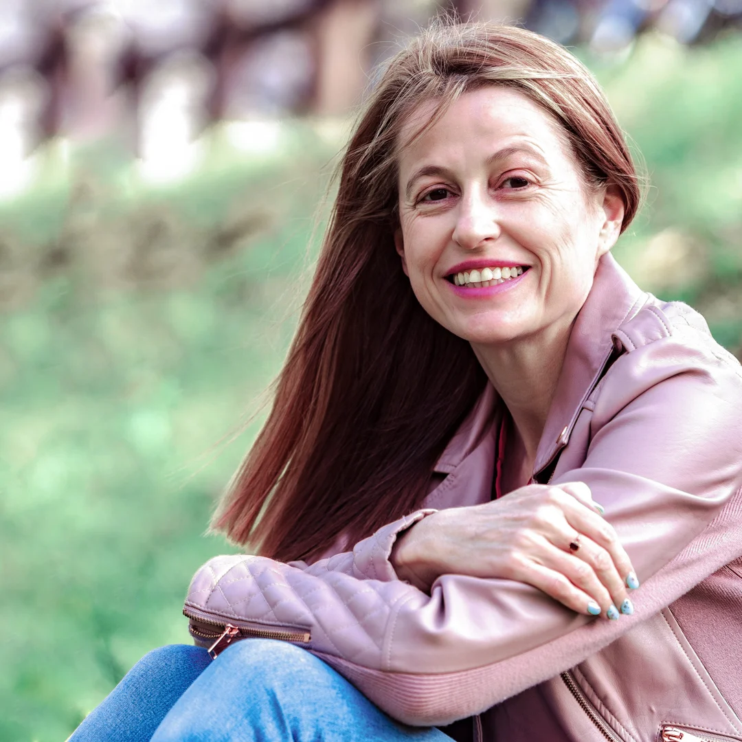 A photo of Japanese Food Guide contributor Louise George Kittaka. She is smiling and sitting side-on on some green grass while turned towards the camera, with her knees bent and her folded arms resting on them. Her hair is long, straight and strawberry blonde. She is wearing blue jeans, a soft pink leather jacket and pink lipstick.