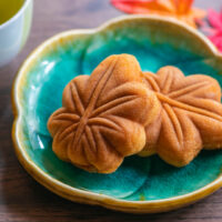 Two maple leaf-shaped momiji manju cakes on a teal ceramic plate with a light brown rim along the scalloped edging. A cup of green tea can partially be seen in the top left-hand corner of the image and two red/orange maple leaves (possibly artificial) at the back-right.
