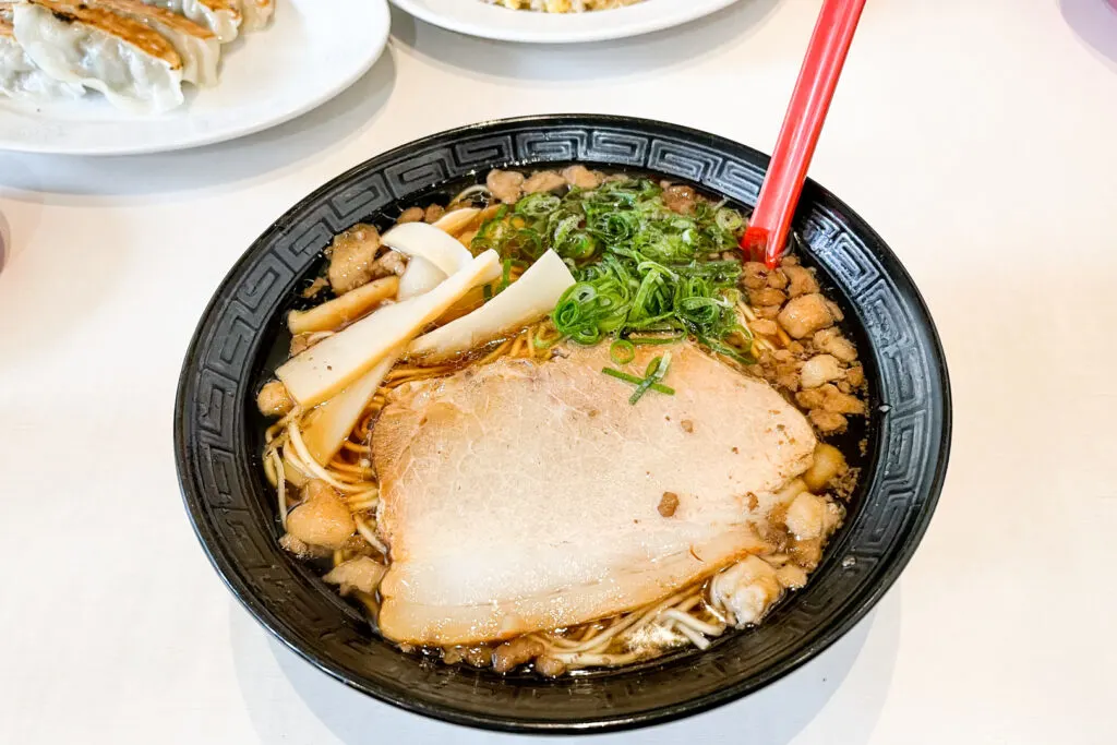 Hiroshima food: Onomichi ramen in a black bowl with a red spoon resting against the side of the bowl. The thin noodles are peeking through the soy-based broth and are topped with chashu (pork slices), menma (dried and fermented bamboo shoots) and scallions (spring onions).