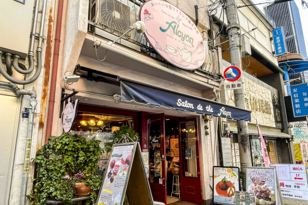 The European-style facade of the Salon De The Alcyon tea shop and patisserie on Dotonbori, Osaka. There are some pot plants and various signboards on the pavement showcasing current offerings.