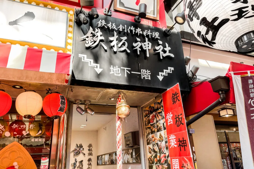 The red, black and white traditional facade of Teppanjinjya teppanyaki restaurant on Dotonbori, Osaka. There is a large white lantern outside and a rope with a bell, that you'd typically see at a shrine, before the stairs leading down to the basement restaurant.