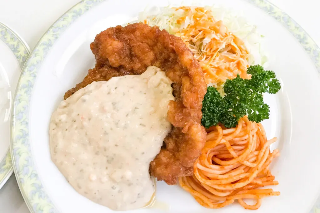 Miyazaki food: Chicken Namban from 'Aji no Ogura Honten' in Miyazaki. The fried chicken thigh is topped with a generous amount of tartar sauce and sits on a bed of shredded cabbage with dressing and a tomato-based spaghetti with a sprig of parsley as a garnish.