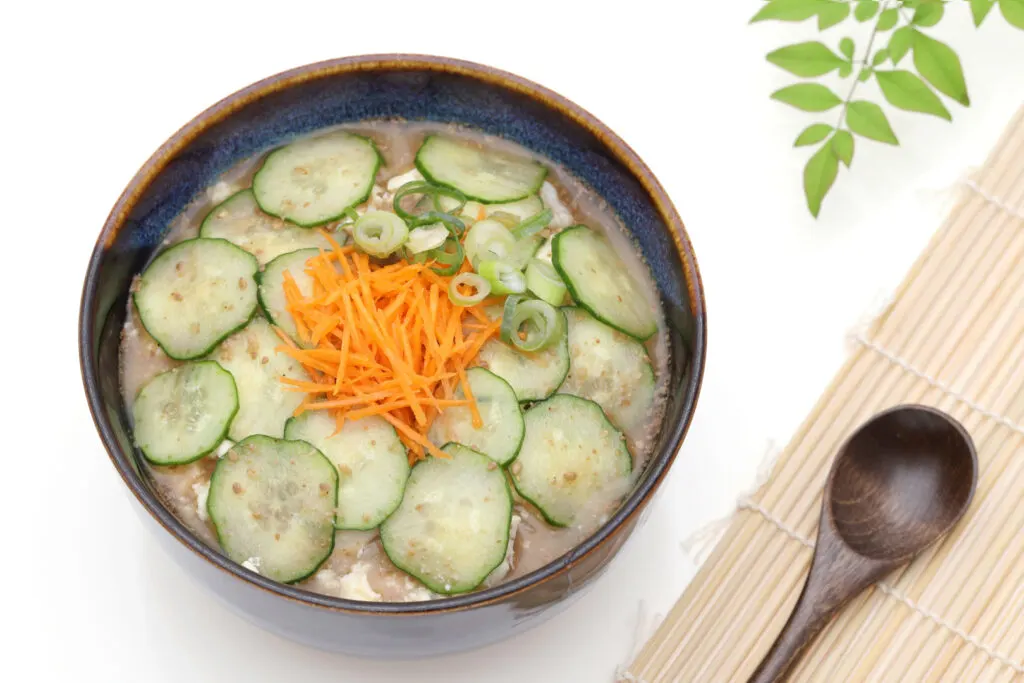 Miyazaki food: A bowl of hiyajiru chilled soup. Sliced cucumbers can be seen carefully arranged on top covering the entire bowl. On top of the cucumber is thinly sliced carrot piled up in the middle along with some sliced green onions (spring onions). To the right of the bowl is a bamboo placemat with a dark wooden spoon on it. Some green foliage also sits on the white surface on the top right of the image.