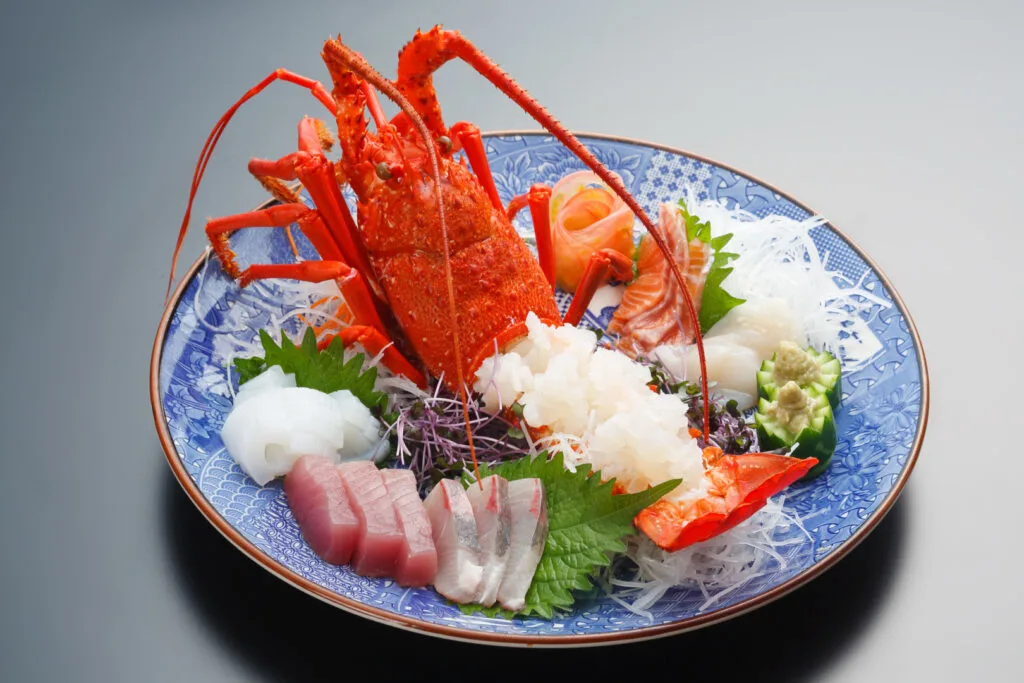 Miyazaki food: A bright red-orange spiny lobster on a blue and white ceramic plate surrounded by sashimi.