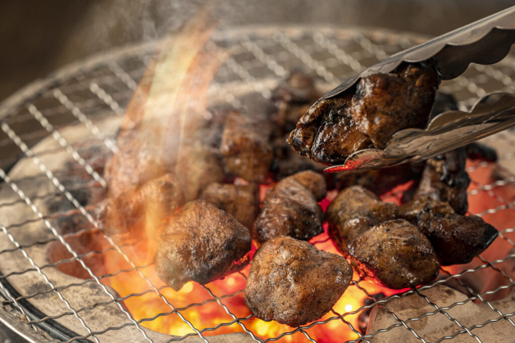 Miyazaki food: Miyazaki's famous chicken, Miyazaki Jidori or Miyazaki Jitokko, being cooked over a small round charcoal grill. Metal tongs from the right side of the image are holding a piece of the chicken. Flames can be seen rising up above the metal grill.