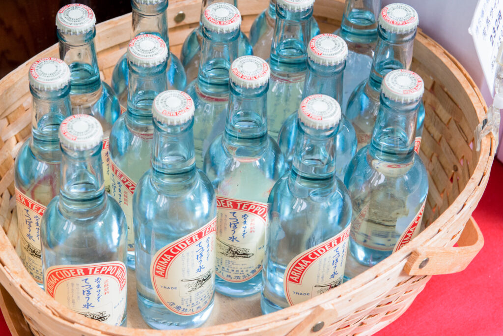 A bamboo basket filled with old-school glass bottles of Arima Cider Teppo Water. The bottles are a light blue color with metal caps and a predominantly red and white label (with some blue and black).