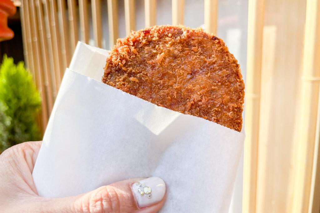 The author is holding a croquette out in front of the camera. The croquette is peeking out of a small white paper bag. Her manicured thumbnail (white nail polish with diamante details) can be seen gripping the front of the bag.
