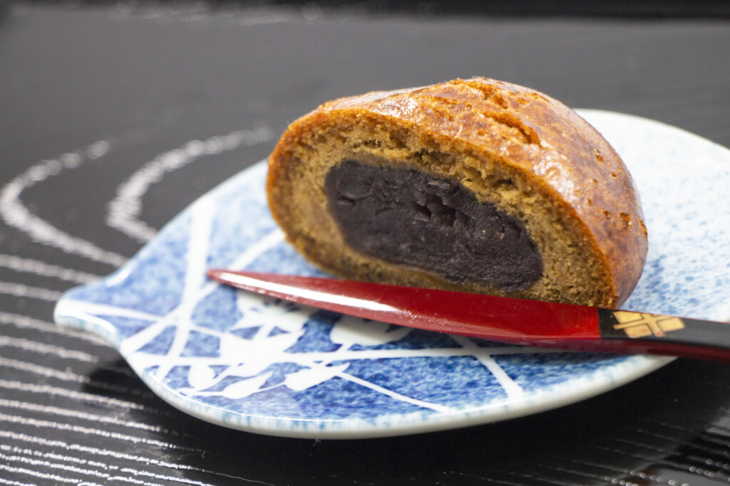 A 'karinto manju' bun cut in half to show the sweet red bean paste inside. The outside is a lovely golden brown color from the brown sugar used to make it. It sits on a blue and white ceramic plate with a red lacquered wooden knife with black and gold detail. The plate sits on a dark wooden surface with a deep and distinctive grain.  