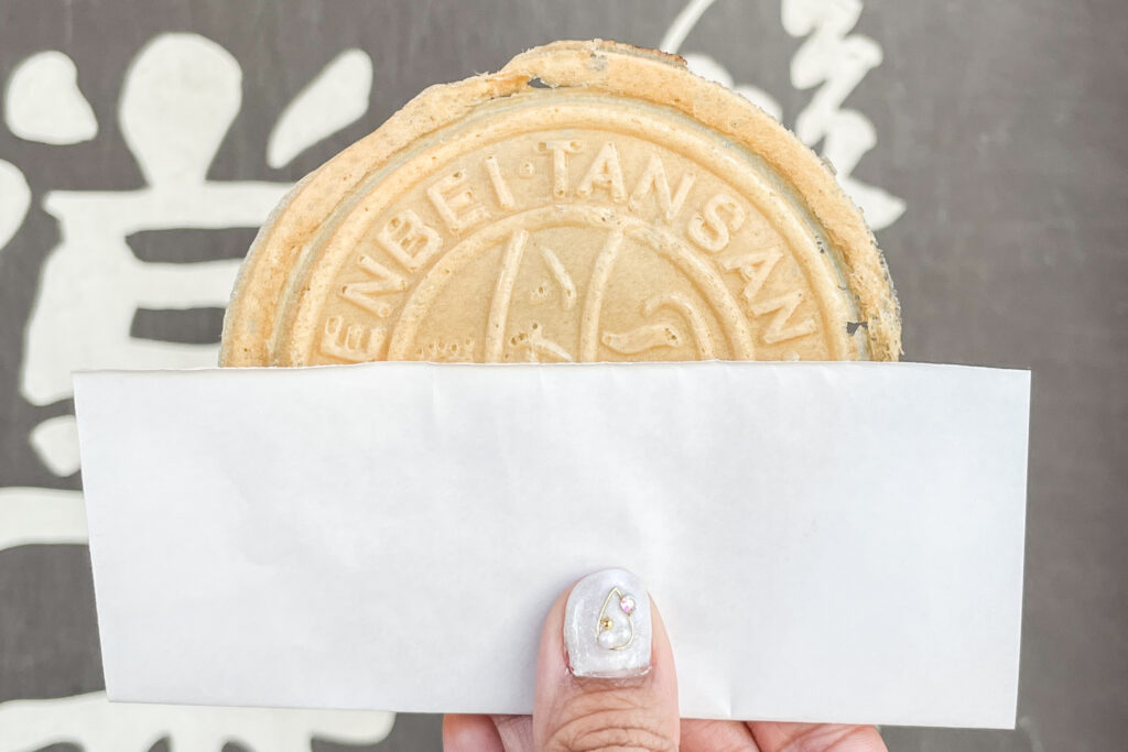 The author is holding a Rare Tansan Senbei rice cracker out in front of the camera. A small piece of white paper is wrapped around the base for easy eating. Her manicured thumbnail (white nail polish with diamante details) can also be seen. 