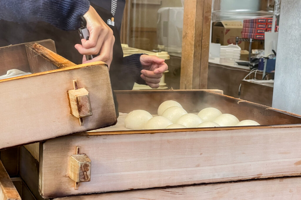 The torso and hands of a person in a traditional navy blue jacket can be seen in frame. The person is picking up one of the steamed buns with metal tongs from a large multi-layered wooden steamer.
