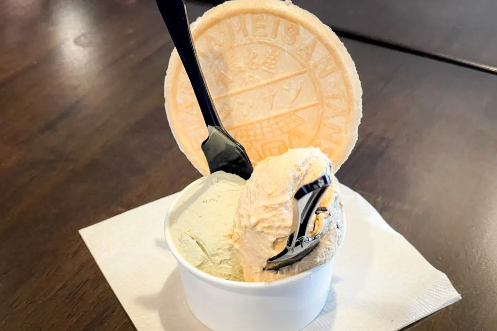Two scoops of ice cream in a cup. One is light green (pistachio) and the other a light caramel (butter caramel flavor). There is a plain Tansan Senbei rice cracker wedged in the ice cream and two black plastic spoons. The cup sits on a white napkin on a dark wooden surface.