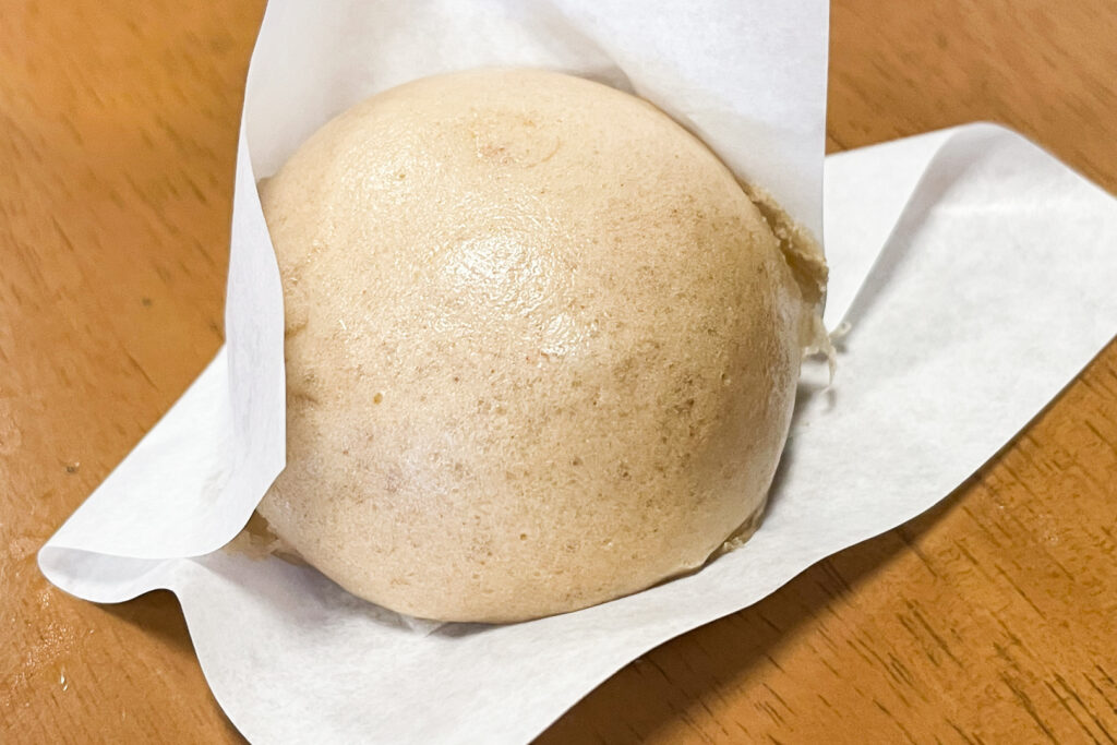A 'yoito manju' steamed bun made from dough containing brown sugar and yam, giving it a bit of a brown-ish tinge. It sits in some paper for easy eating and has been placed on a wooden surface for the photo.