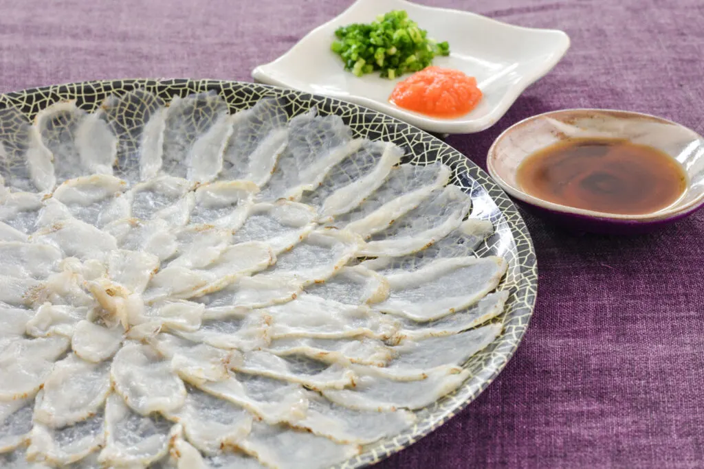 Yamaguchi food: Very thinly-sliced fugu sashimi beautifully arranged on a large plate. A small bowl of dipping sauce and a side dish with other condiments sit beside it, all placed on a purple tablecloth.