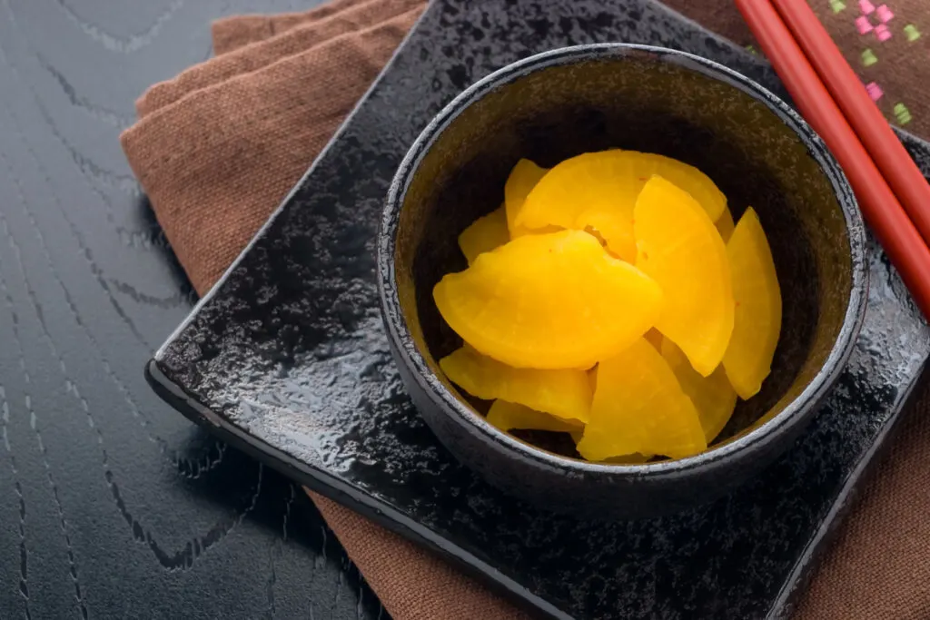 A small, dark ceramic bowl partially filled with pickled daikon radish (takuan) roughly cut into half-moon shapes. The yellow color of the takuan contrasts nicely with the dark crockery. The bowl sits on a matching square plate and a brown placemat. Red chopsticks can be seen resting on the side of the plate. Everything sits on a dark wooden surface.