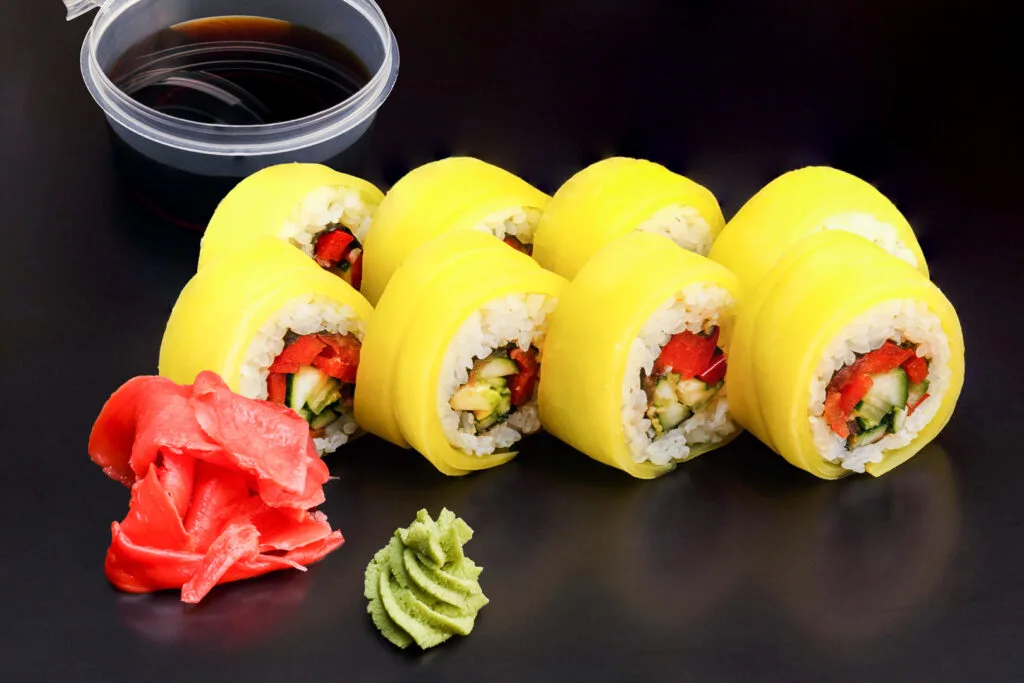 Strips of pickled daikon radish wrapped around sushi rolls (imagine yellow pickled daikon instead of dark nori seaweed). There are eight pieces of sushi and the bright fillings can be seen in the center of each piece. In front of the sushi is some red pickled ginger and wasabi, and behind a small, open container of soy sauce.