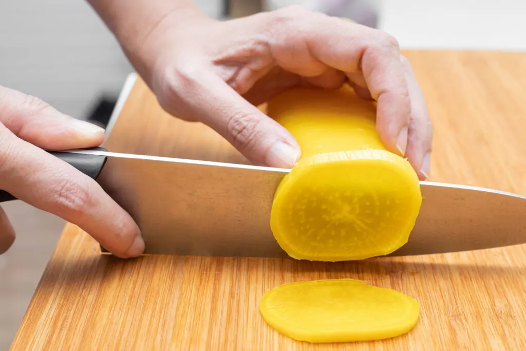 A whole pickled daikon radish being cut into slices on a wooden chopping board with a large knife. One hand of the person cutting is holding the radish steady while the other is holding the knife and slicing through the takuan.