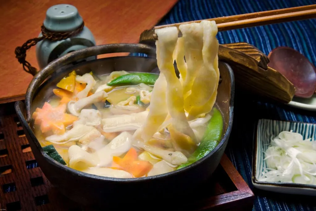 Yamanashi food: A metal hot pot filled with 'hoto' noodle soup sits amongst a table setting. Chopsticks are coming into frame from the right and are holding up several wide, flat strands of noodles, while various vegetables can be seen floating in the broth.