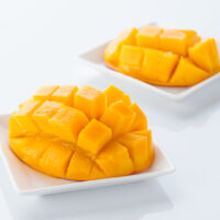 Two slices of Miyazaki mango still attached to the skin sitting on white square plates. The flesh has been cut both vertically and horizontally, and then the skin inverted so that the pieces of still-attached mango stand up in little cubes.