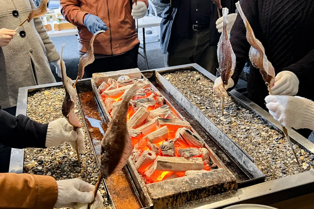 Matsukawa-ura Lagoon, Soma City: Four people wearing white gloves place their skewered fish into the shell-filled sand on the beach BBQ, facing towards the burning charcoal in the center.