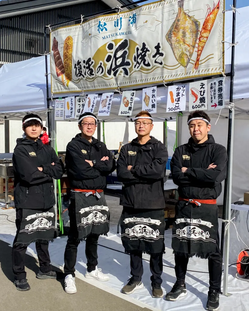 Four members of Matsukawa-ura Team Guide stand with arms crossed in a very "Iron Chef-like" pose in front of a hamayaki beach BBQ set-up in Matsukawa-ura, Soma City. They are wearing black clothes, black aprons with white text and white bandanas.