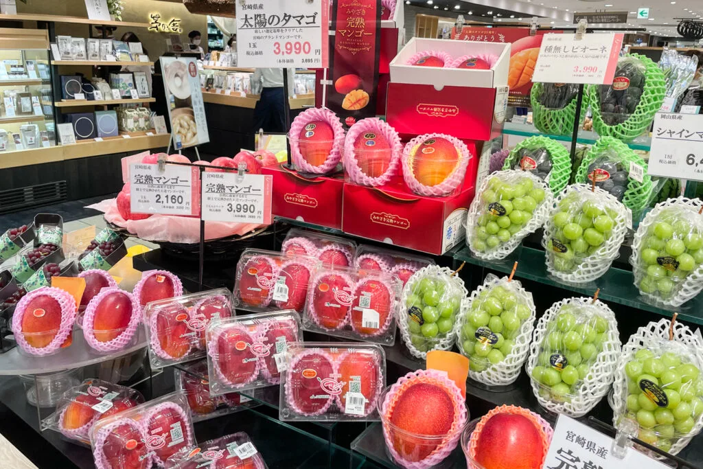 A Japanese supermarket display with Miyazaki mangoes on one side and green grapes on the other. At the top of the mango display are 'Taiyō no Tamago' Miyazaki mangoes (3990 yen each). Under them there are regular Miyazaki mangoes - 3990 yen for two or 2160 yen for one.