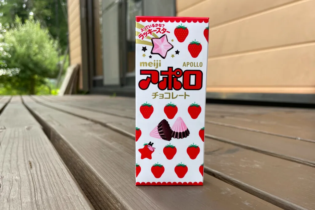 A box of Apollo Chocolates. There is imagery of the snack, like a little ridged mountain peak with pink chocolate at the top, and a regular darker chocolate color at the base. There is also imagery of strawberries and stars on the white, red and pink colored packaging.