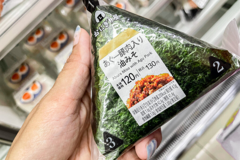 Okinawa food: The author's hand can be seen holding up an onigiri (rice ball) to camera at a convenience store in Okinawa. The onigiri's flavor is 'Abura Miso and Agu Pork'.