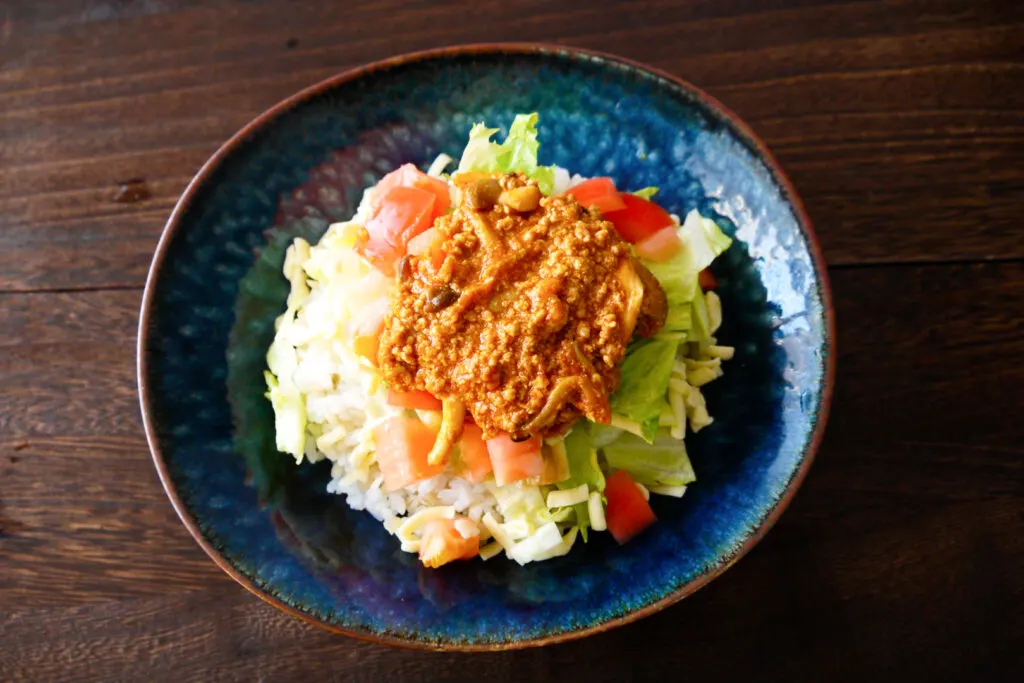 Okinawa food: An Okinawan dish called 'Taco Rice' on a dark blue ceramic plate sitting on a dark wooden surface. It is white rice topped with lettuce, tomato and cheese, and then a meat taco mix (like having taco ingredients on rice instead of in a shell).