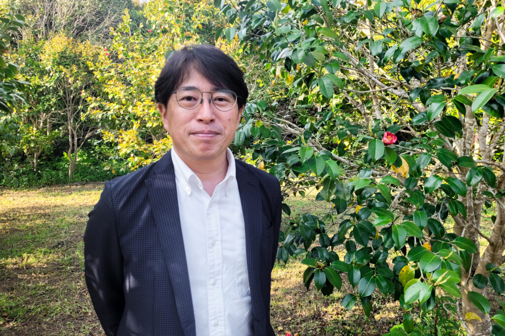 MTG President Tomitaka Tanigawa standing beside a camellia tree (with one red flower in bloom) at Tsubaki no Goto's camellia garden on the Goto Islands. He wears a white business shirt and black suit jacket. He has short black hair and glasses, and is smiling without his teeth showing.