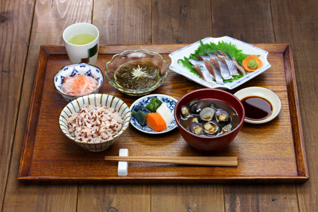 A wooden tray with a traditional Japanese meal set out on it. At the front-left is a bowl of multi-grain rice and the front-right a bowl of clam soup. At the top-right is the main dish (fish). There are two side dishes (seaweed and glass noodles), and some pickles. There is also a cup of green tea at the back-left, and chopsticks on a chopstick rest at the very front.