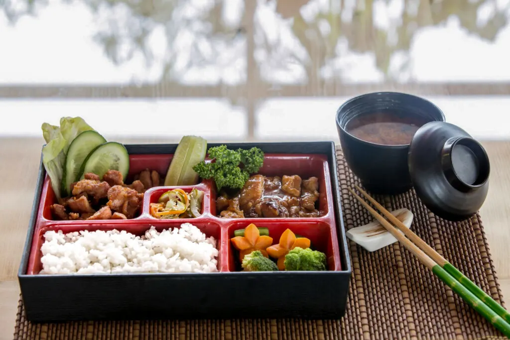 A Japanese bento box with the rice placed at the front-left. There is a bowl of miso soup placed on the table to the right of the bento box, along with some chopsticks on a chopstick rest.