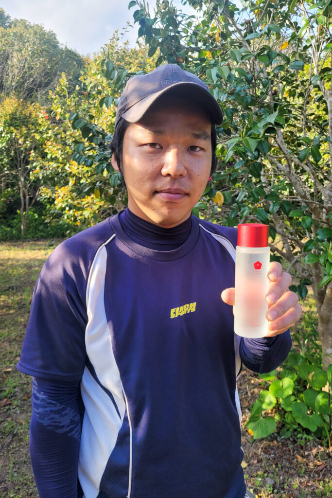 Garden overseer Hayato Imamura stands in the camellia garden of 'Goto no Tsubaki' holding a bottle of camellia oil skin toner made from Goto camellias. The plastic bottle is clear with a red camellia on it and has a matching red lid. Imamura wears navy blue sports clothes and a black baseball cap.