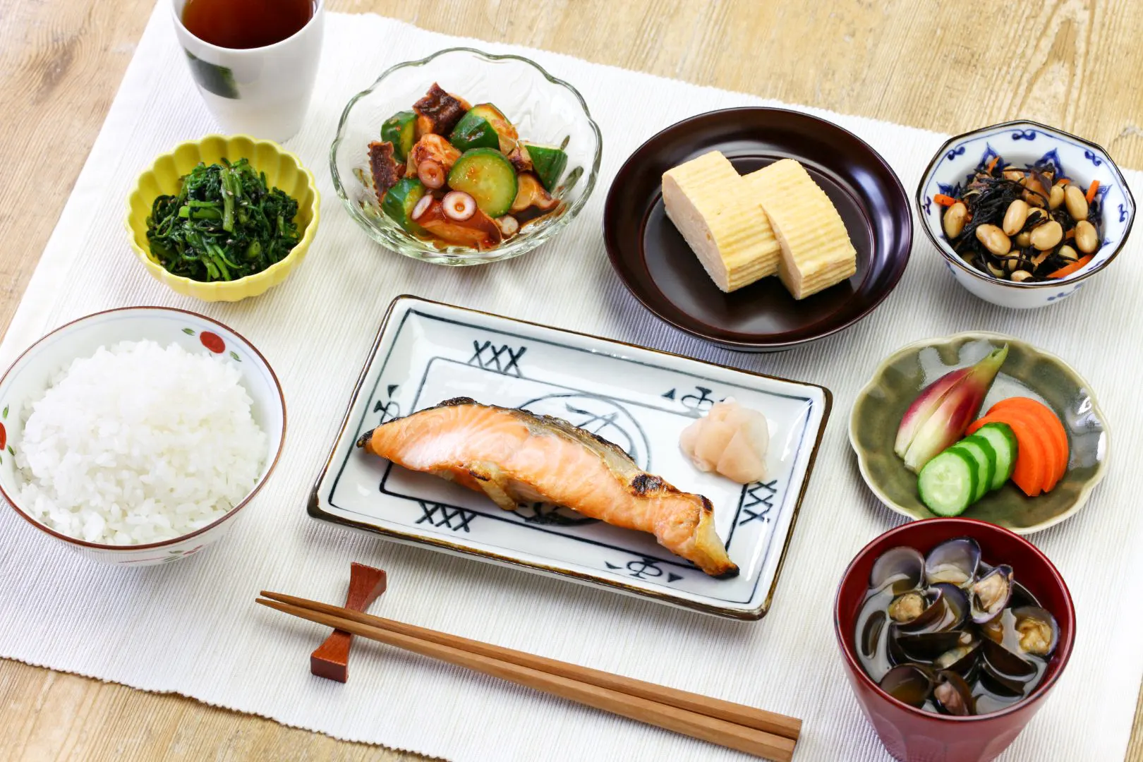 A traditional Japanese meal laid out on a white fabric placemat. There is rice to the left, soup to the right and fish in the center. There are four side dishes plus pickles, and a cup of dark-colored tea. There are wooden chopsticks sitting on a red chopstick rest at the front of the meal, with the tip of the chopsticks facing to the left.