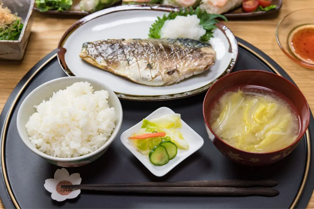 A black, round lacquered tray with rice to the front-left and soup to the front-right (a small dish of pickles can be seen between them). Behind them in the center is the main dish (fish). Slightly in frame, various other side dishes can be seen on the table.